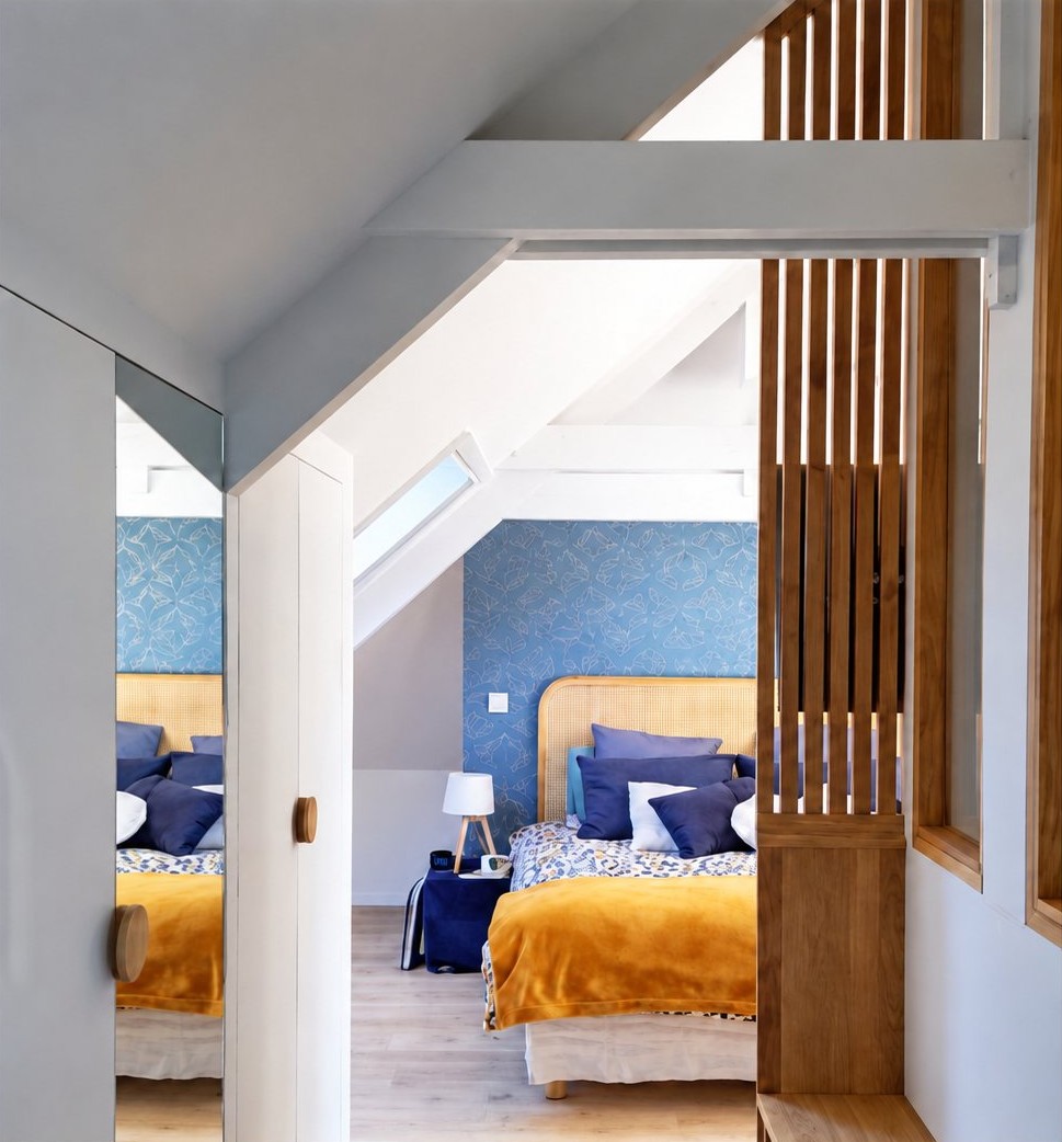 Chambre mansardée aux murs bleus avec lits et plaids moutarde, séparée du couloir par un claustra en bois.