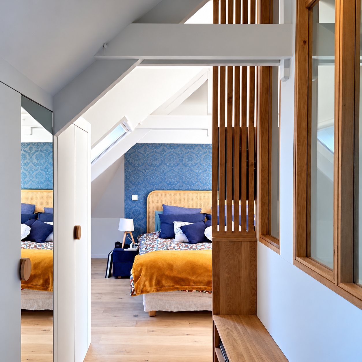 Chambre mansardée aux murs bleus avec lit à tête en rotin et plaid moutarde, derrière une cloison en bois à claire-voie.