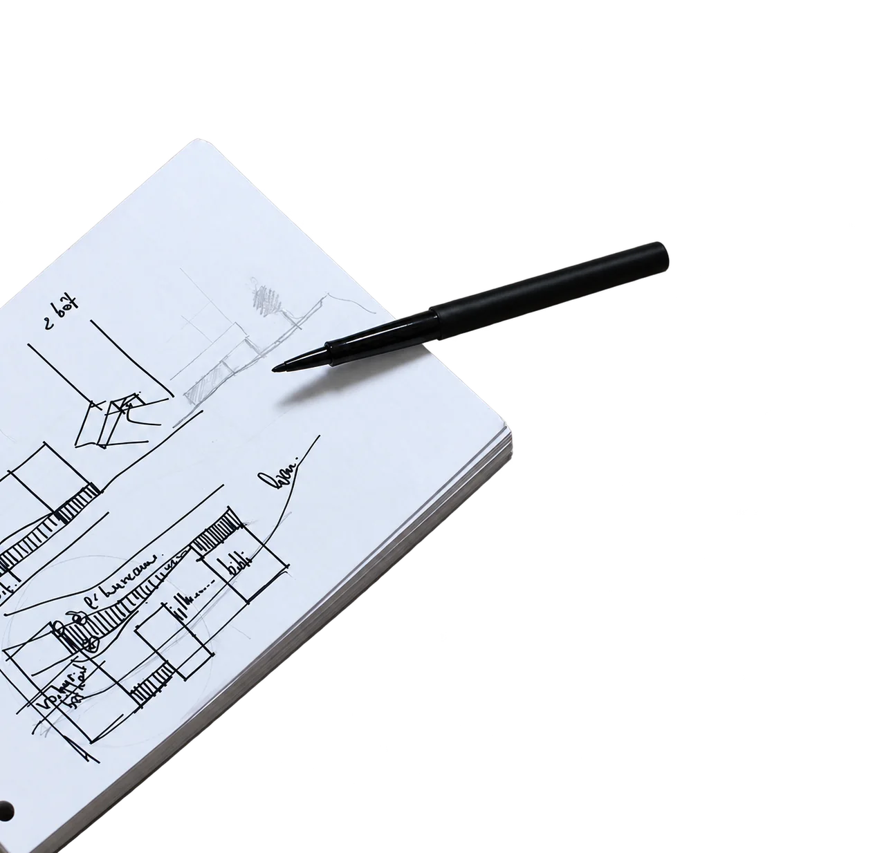 Un carnet ouvert montrant des croquis au feutre noir de plans et de coupes, posé sur un panneau de bois aggloméré avec un stylo noir.