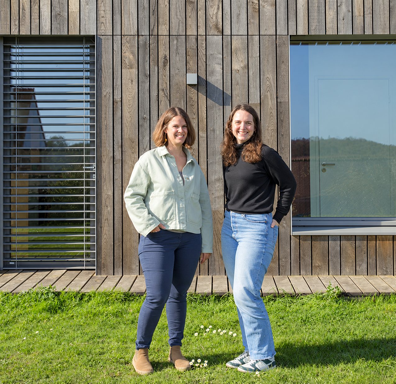 Deux femmes souriantes posent côte à côte sur une pelouse fleurie de pâquerettes, devant une façade en bois avec un brise-soleil et une grande fenêtre.