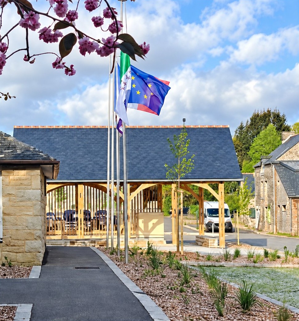 Halle de village ouverte en bois sous un toit d’ardoise, devant des mâts de drapeaux, entourée de pierre et d’un cheminement planté.