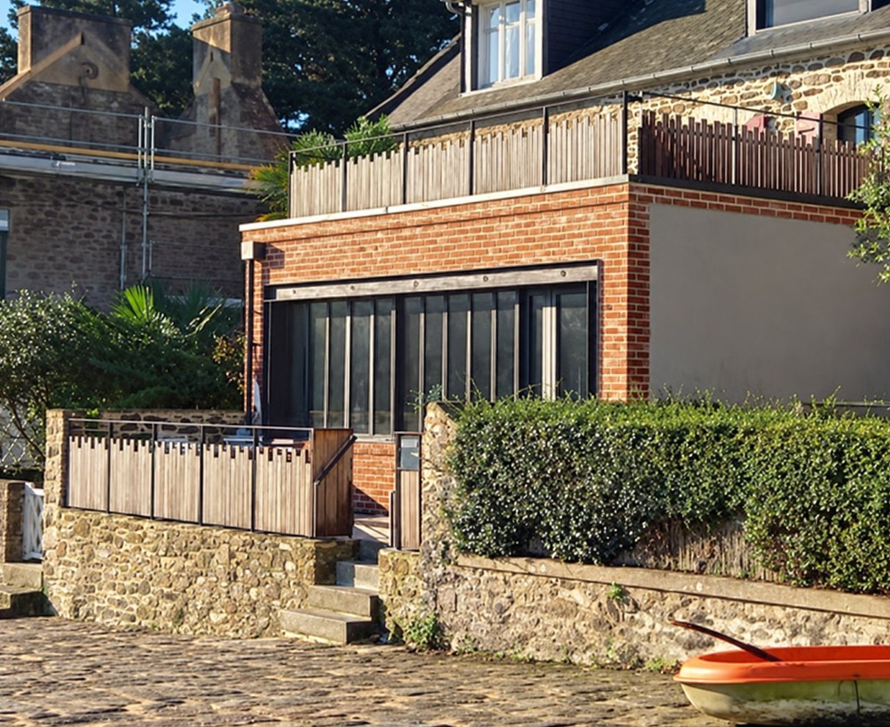 Maison en brique et pierre avec terrasse en bois en bord de Rance, derrière un muret.