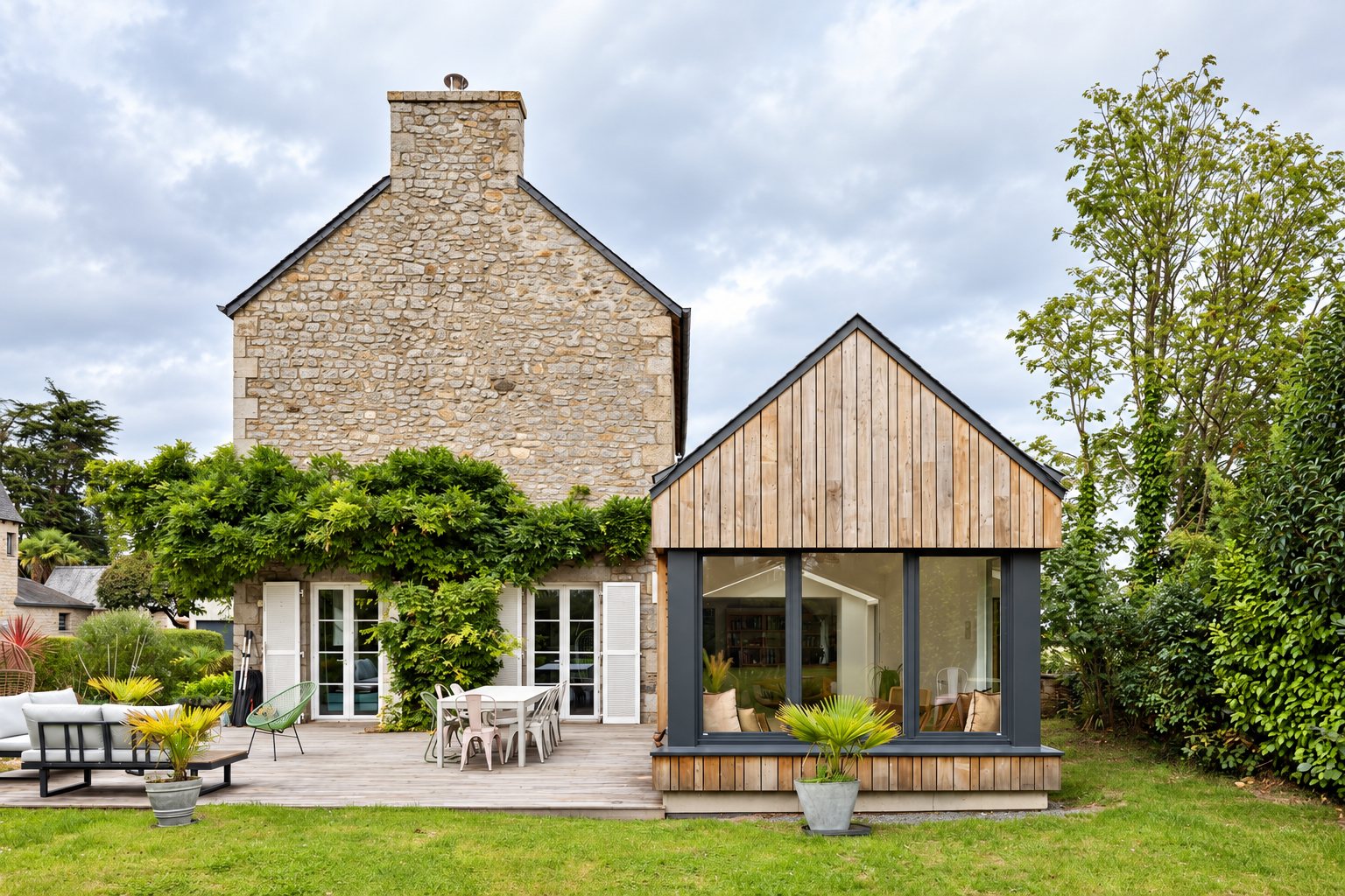 Une maison ancienne en pierre dotée d'une extension en bois à grande baie vitrée, avec une terrasse en bois et un jardin au premier plan.