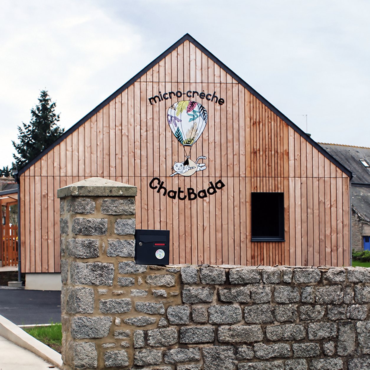 Façade en bois d’une micro-crèche à pignon portant une montgolfière peinte, derrière un muret en pierre.