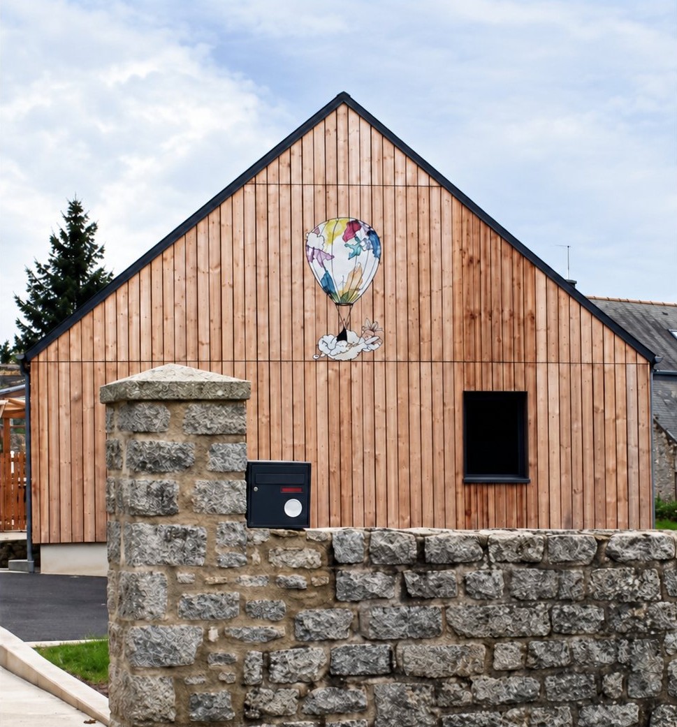 Façade en bois d’une micro-crèche à pignon portant une montgolfière peinte, derrière un muret en pierre.