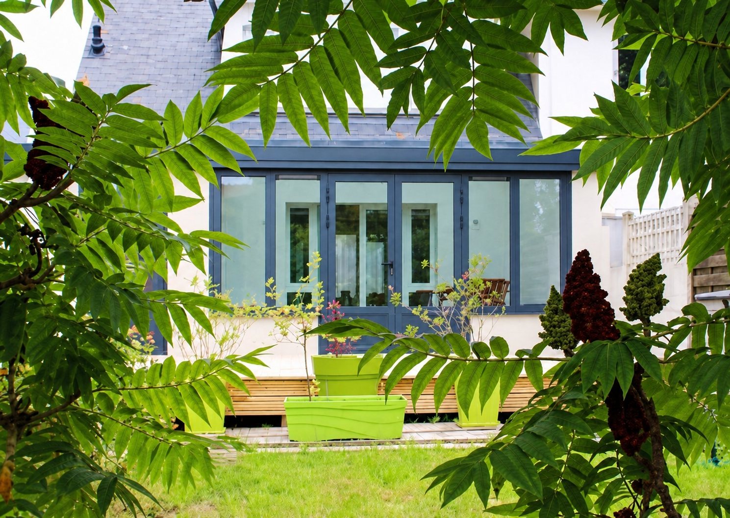 Véranda à grandes baies vitrées donnant sur un jardin planté et une terrasse en bois.