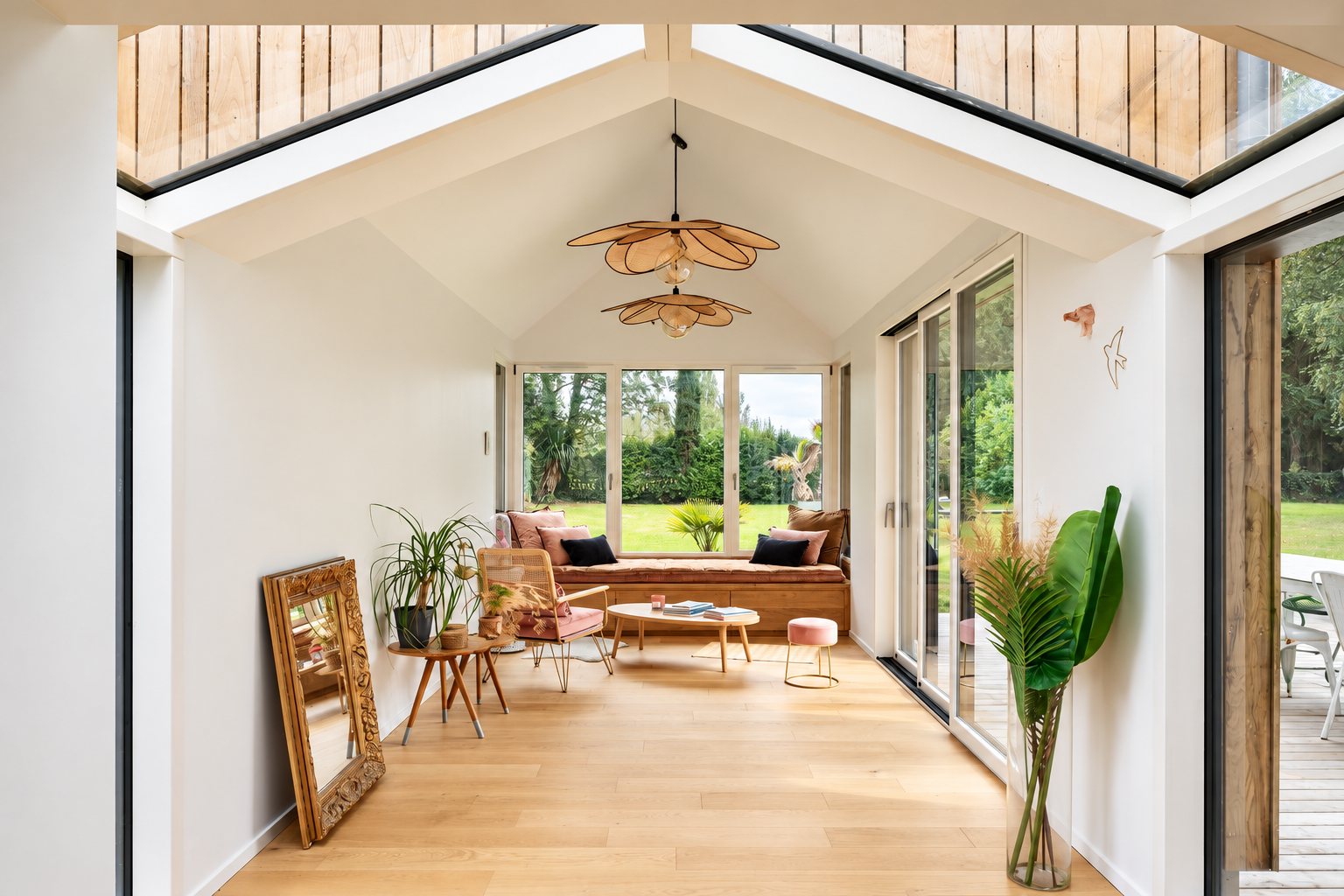 Une véranda moderne aux murs blancs et au parquet clair, meublée d'une banquette sur mesure, d'un fauteuil rose, de suspensions en rotin et d'un grand miroir doré, ouverte sur un jardin.